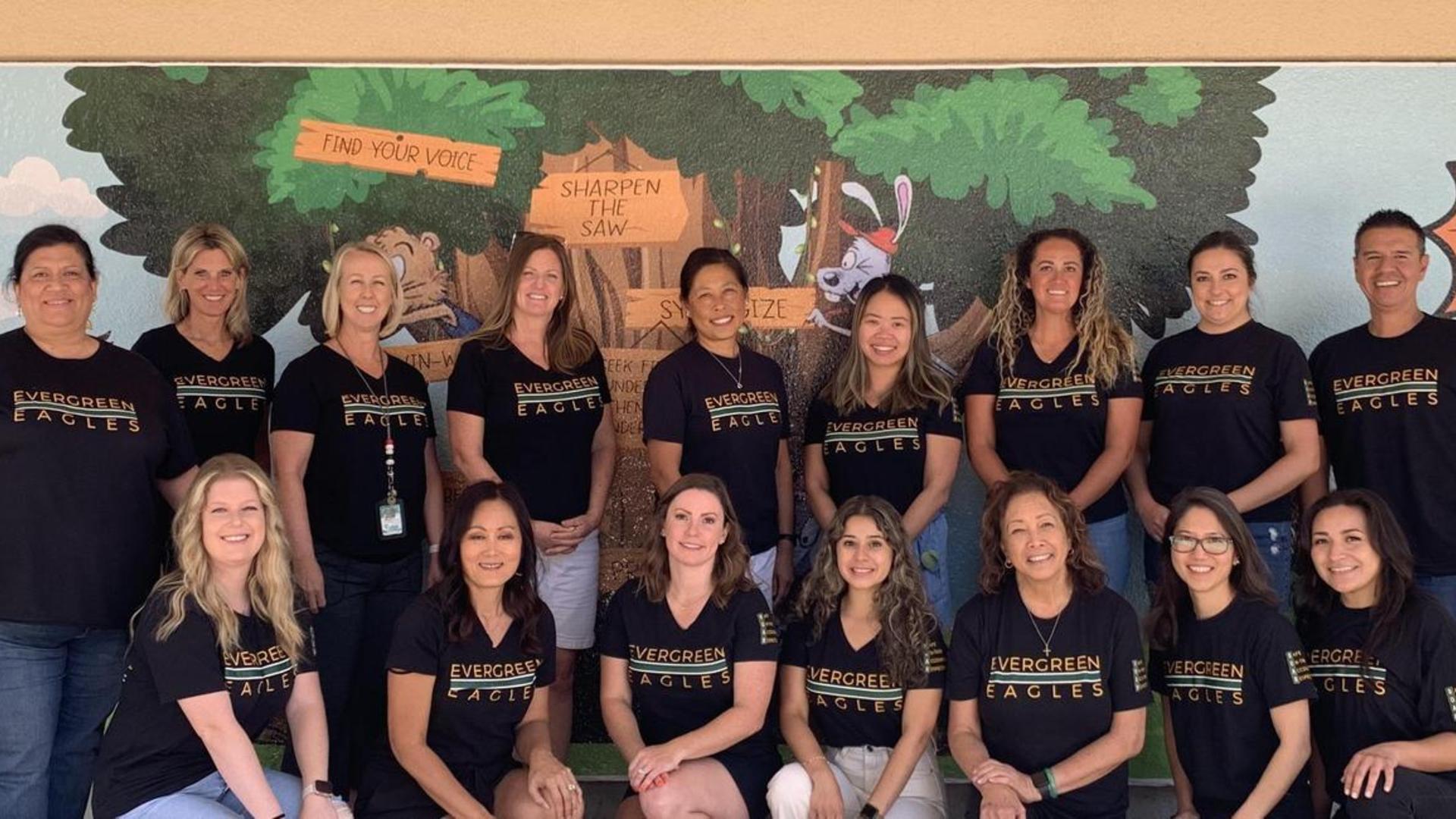 Group of teachers in black shirts smiling in front of a mural.