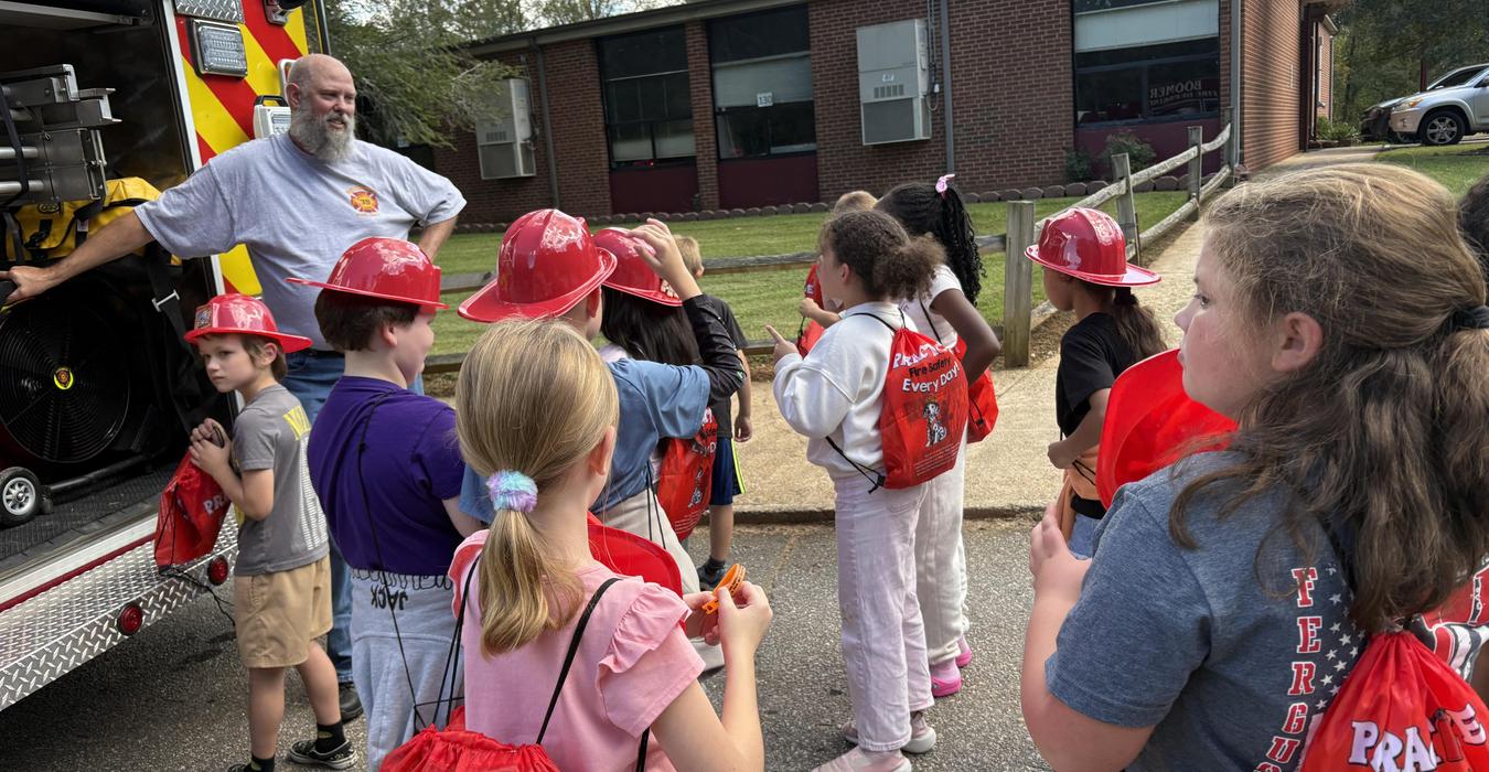 Children in red hats listen to a fireman by a fire truck outside a school.