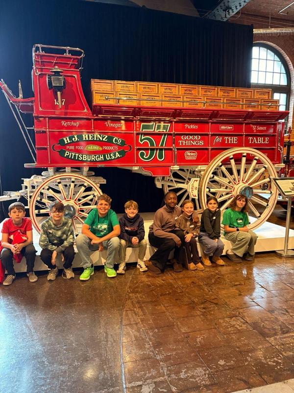 Image depicts a team of Quaker Valley students posing in front of a large, red wagon at the Heinz History Center.