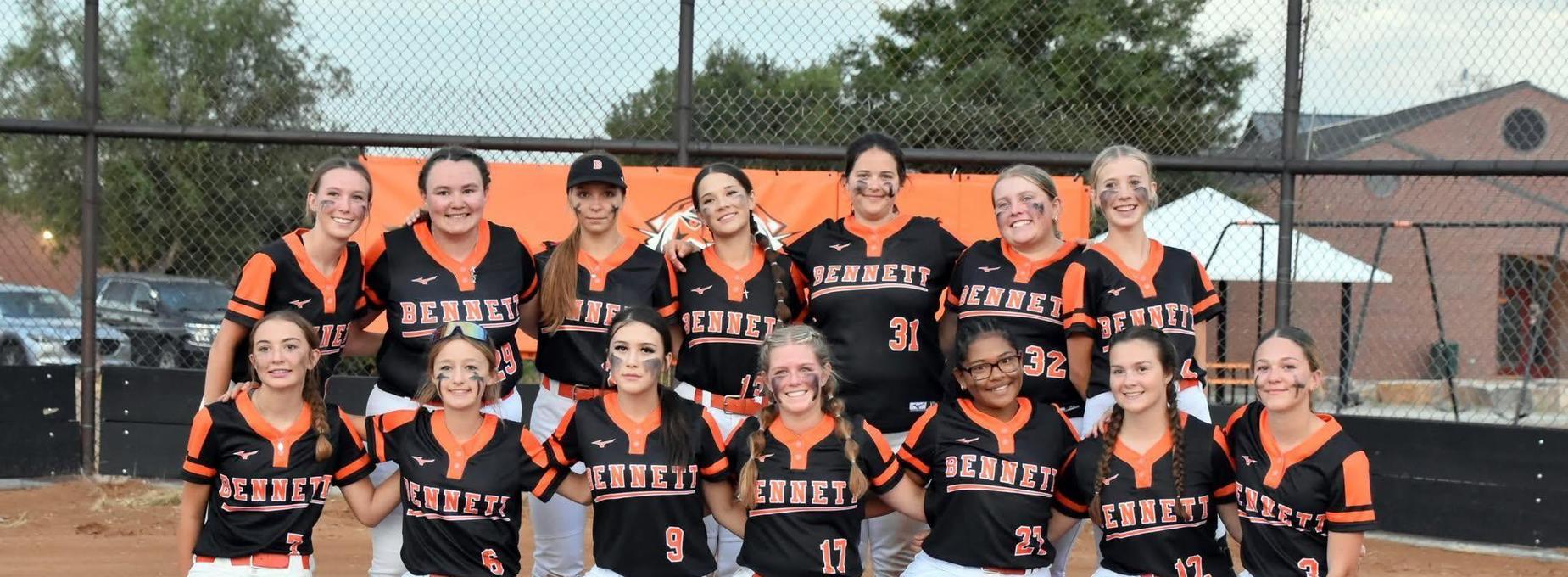 Group of teenage girls in orange and black softball uniforms posing in front of a field.