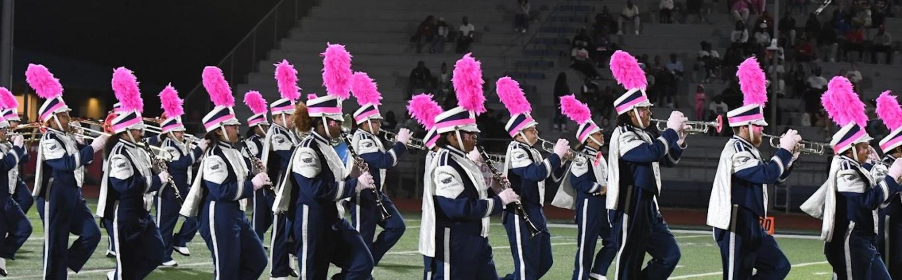 Marching band members in uniforms with pink feathered hats performing during a halftime show.