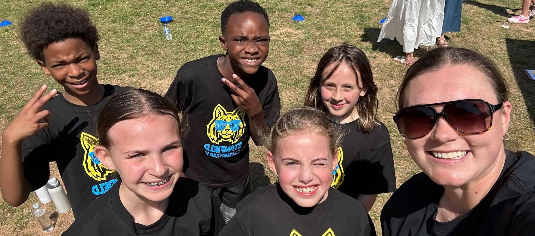 Group of children and an adult, all smiling, wearing matching black shirts outdoors.