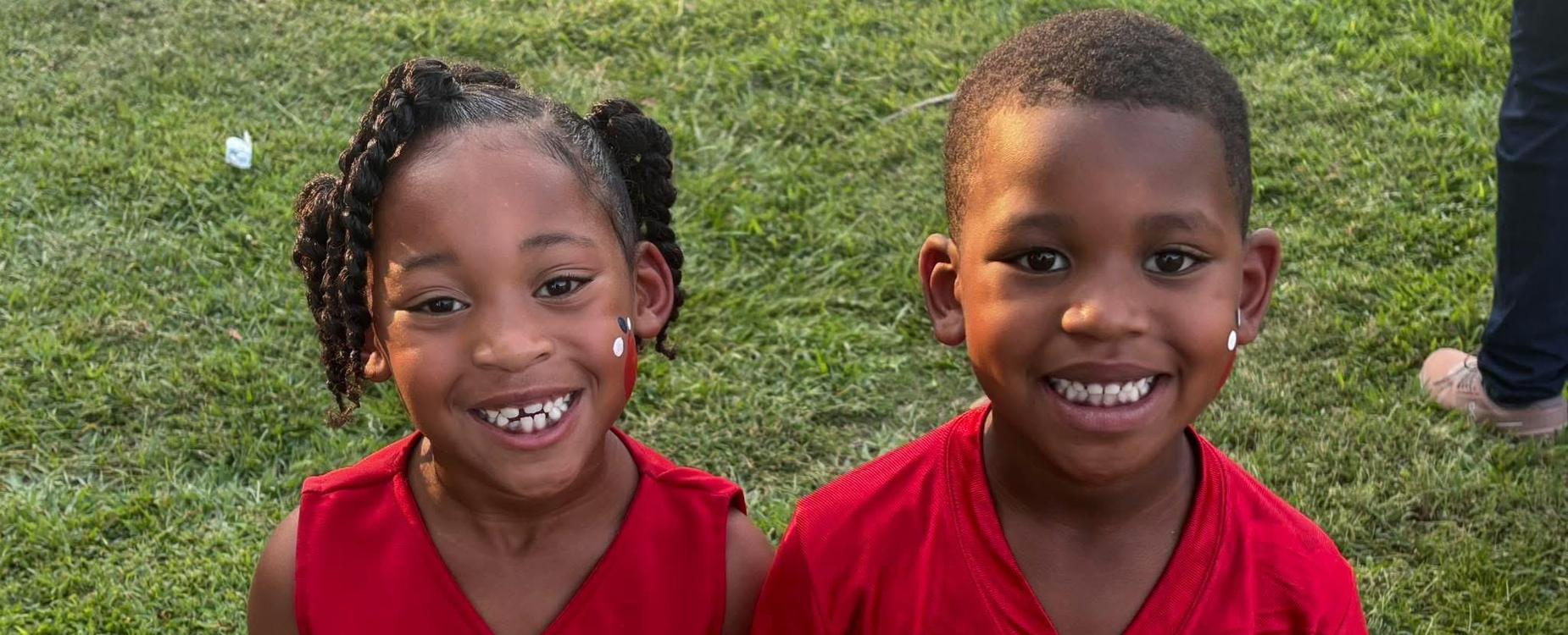 Two smiling children in sports jerseys standing on grass.