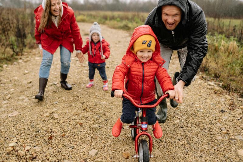 Two adults cheer on a young child riding a red bike on a gravel road.