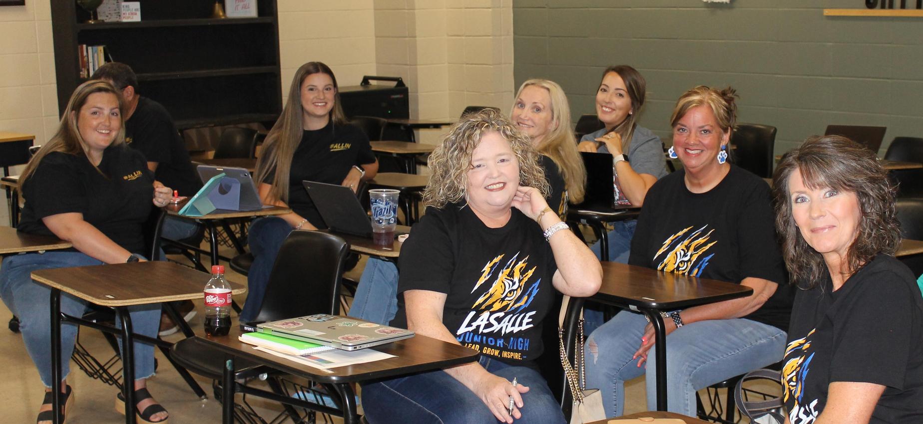 A group of smiling educators seated in a classroom, wearing matching black shirts.