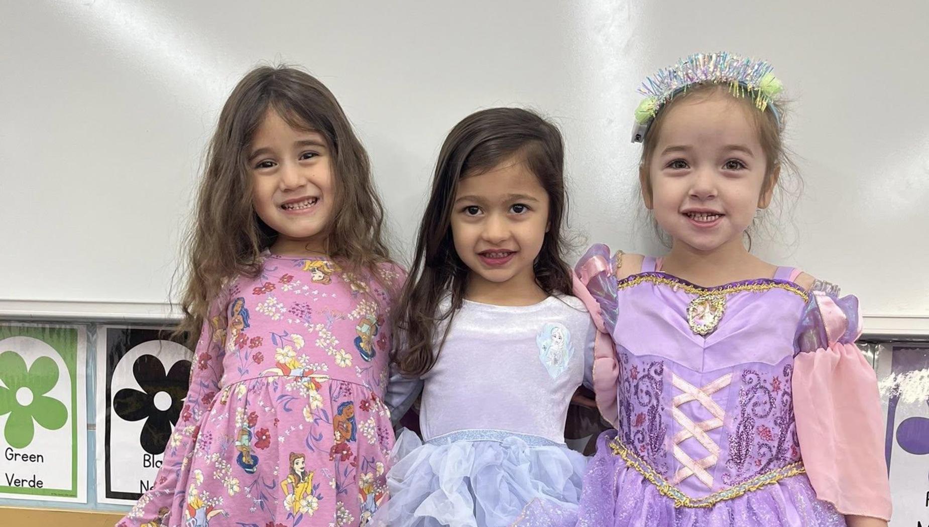 Three girls in colorful dresses pose together in a classroom setting.