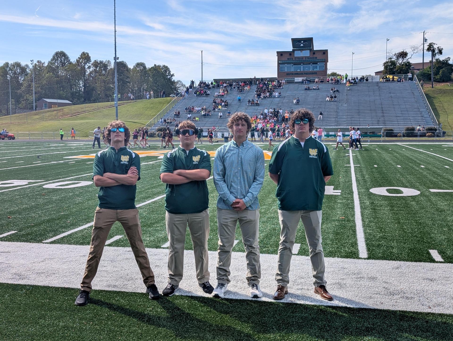 Four students in matching uniforms standing confidently on a sports field with spectators behind.