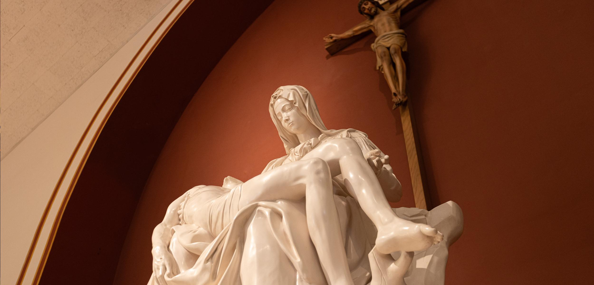 Marble statue of Mary holding Jesus, with a crucifix in the background.