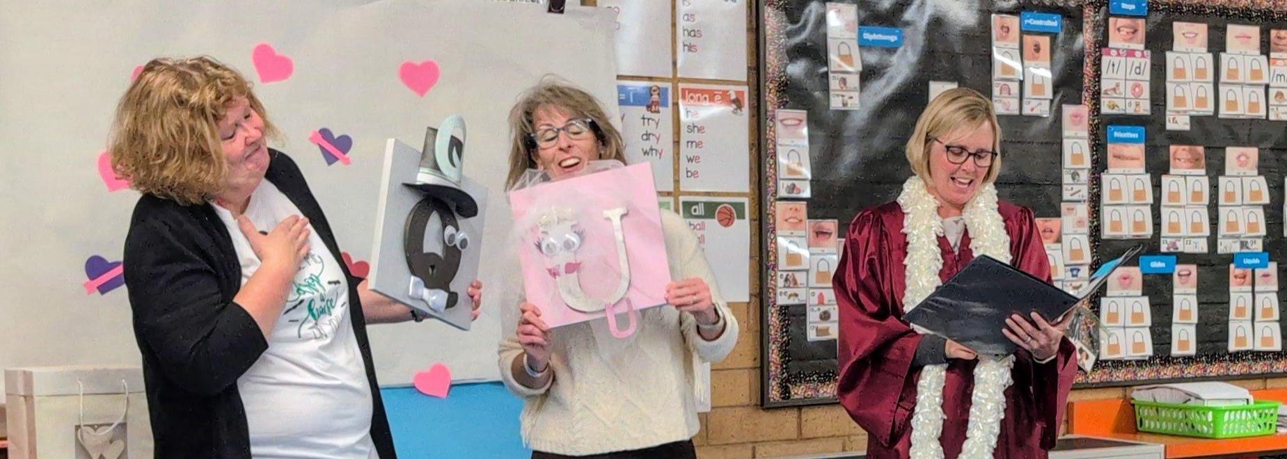 Three women in a classroom, one holds a paper cutout, another holds a sign, and the third reads from a booklet.