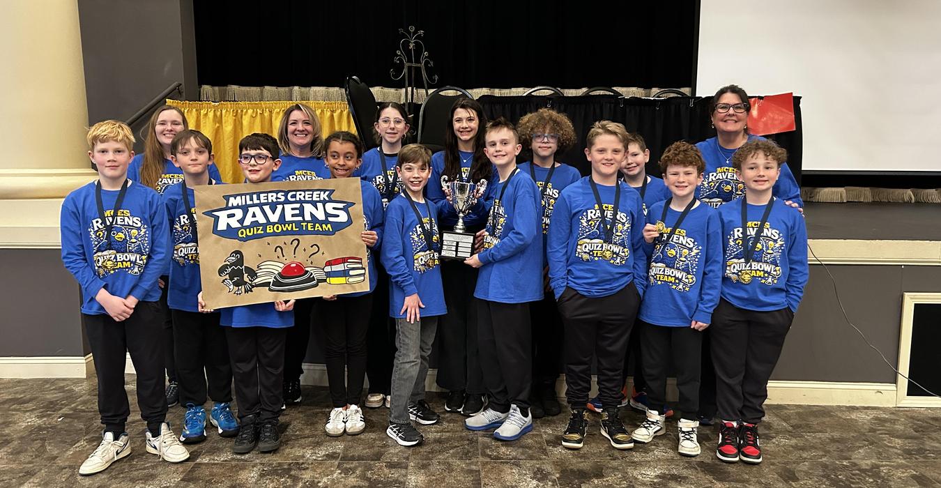 Team of children in blue shirts holding trophies in a school auditorium.