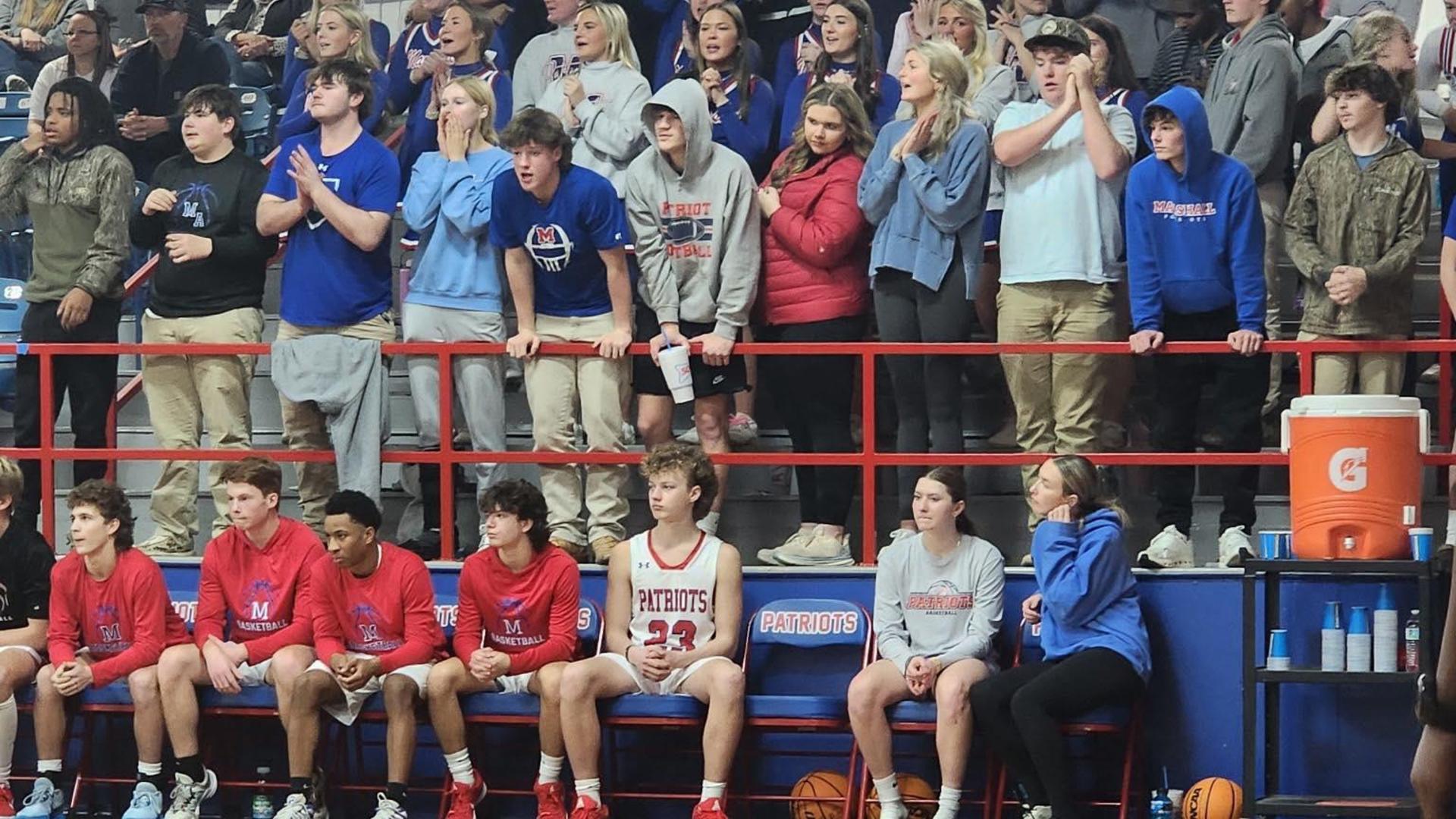Fans and teammates watch a basketball game from the stands, showing various emotions.