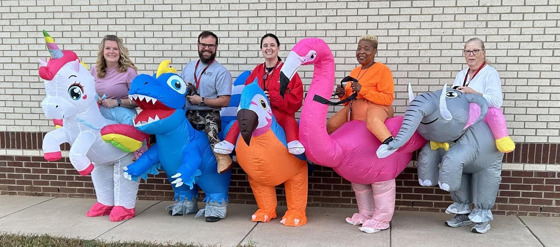 Group of people in inflatable dinosaur and animal costumes standing against a wall.