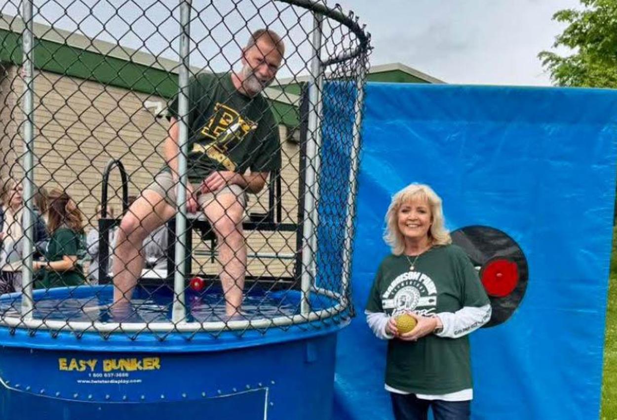 Mr. Marasti sits in a dunking booth and Mrs. Socrates stands next to the booth holding a yellow ball