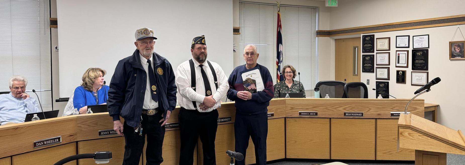Three men at a meeting, one holding a certificate, with attendees in the background.