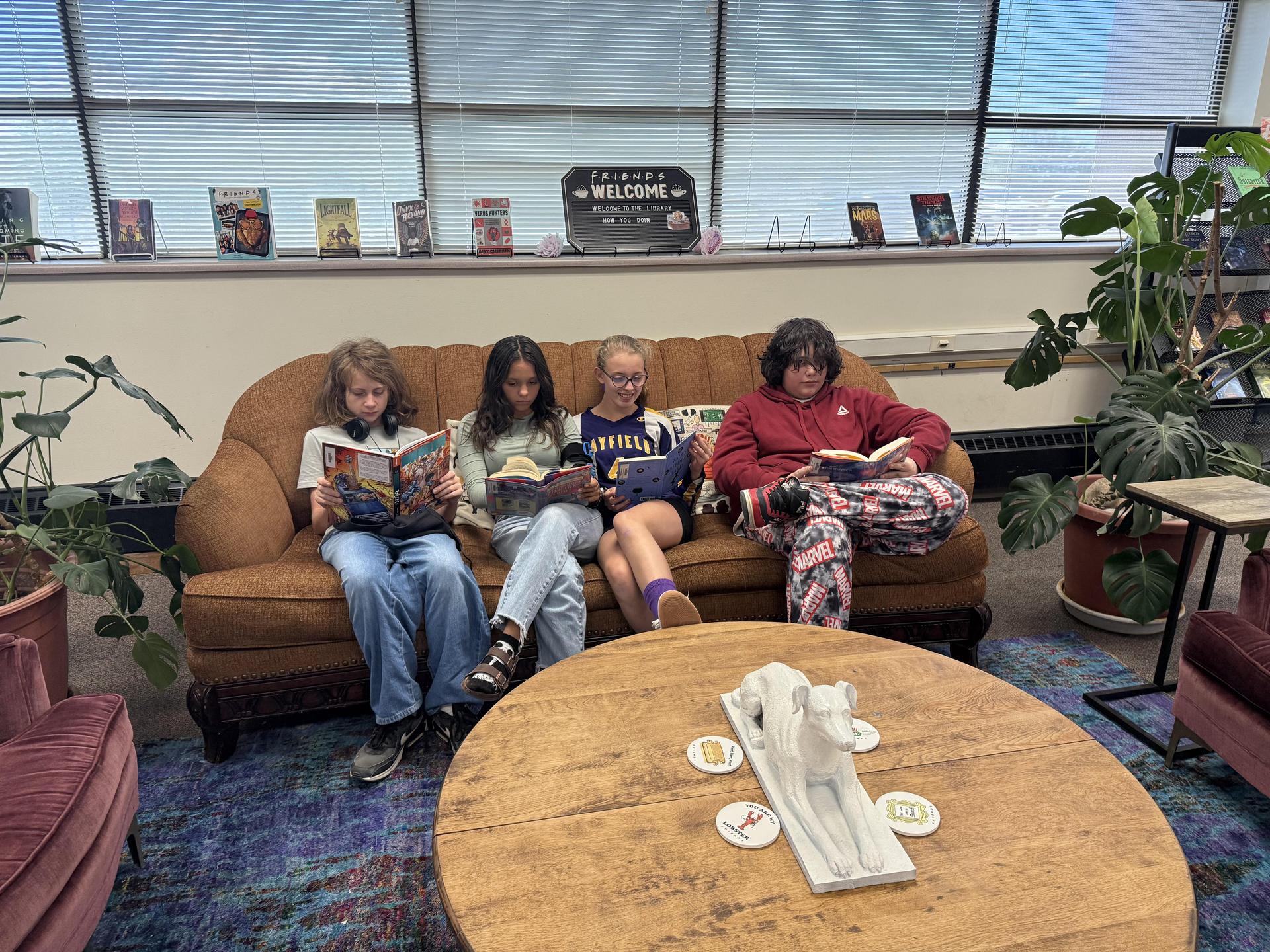 Three children reading books on a couch surrounded by plants and a coffee table.
