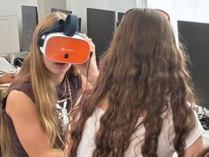 A person wears an orange virtual reality headset while sitting next to another student in a classroom setting.