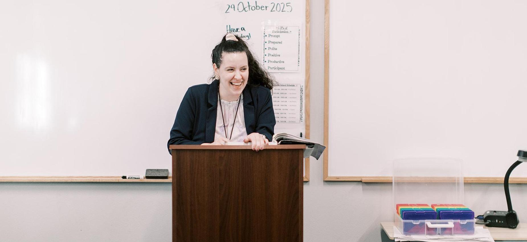 Teacher smiles from a podium at the front of the classroom