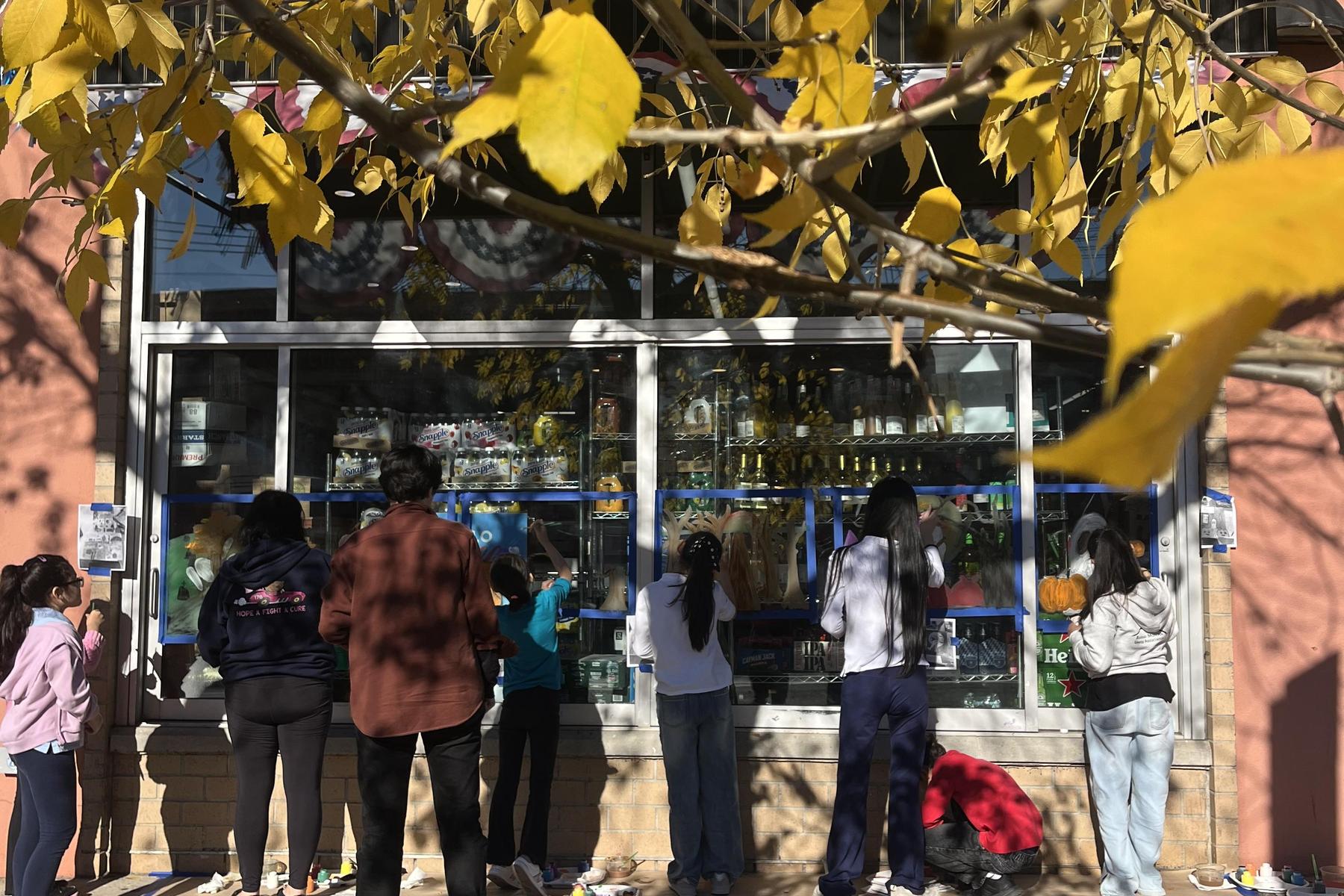 Participants painting a shop window while surrounded by autumn foliage.