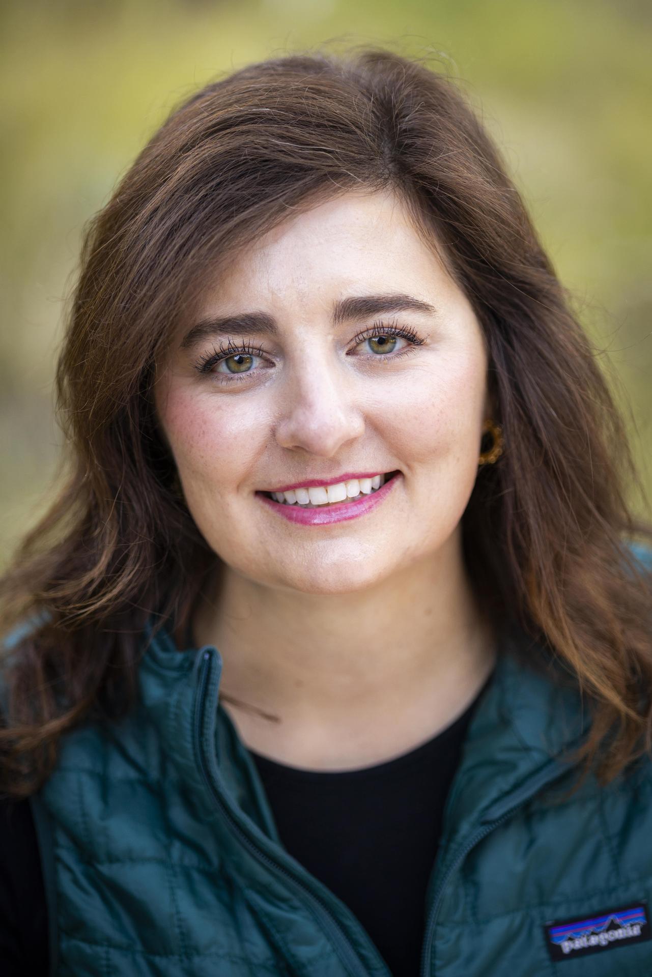 A smiling woman with brown hair wearing a green vest, outdoors.