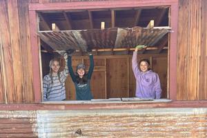 Three middle school students wearing gardening gloves smile from inside a shed