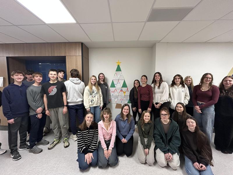 A group of students posing in front of a decorated Christmas tree on a wall.