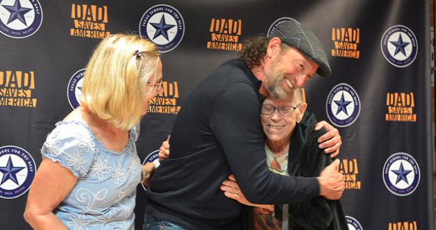 Academy Award-winning actor Troy Kotsur shares a heartfelt hug with one of his biggest fans at an event. Both are smiling warmly, radiating joy. A woman stands beside them, also smiling. The backdrop features the Texas School for the Deaf logo and the text 'Dad Saves America,' adding context to the celebratory moment.