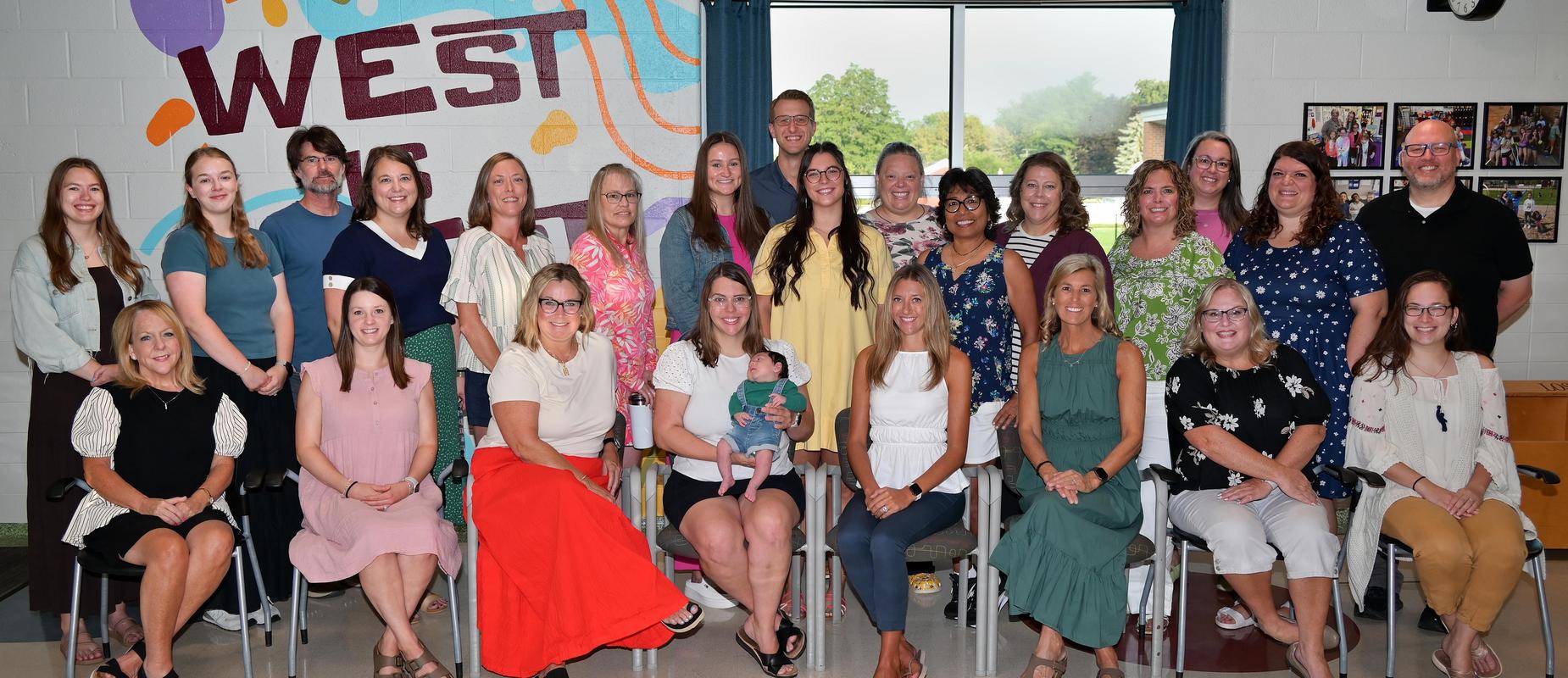West staff pose inside their building foyer in two rows with a teacher holding her baby in the center