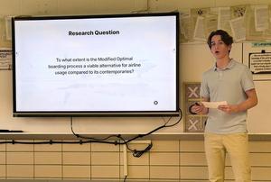 A student presents a research question about airline boarding processes on a screen.