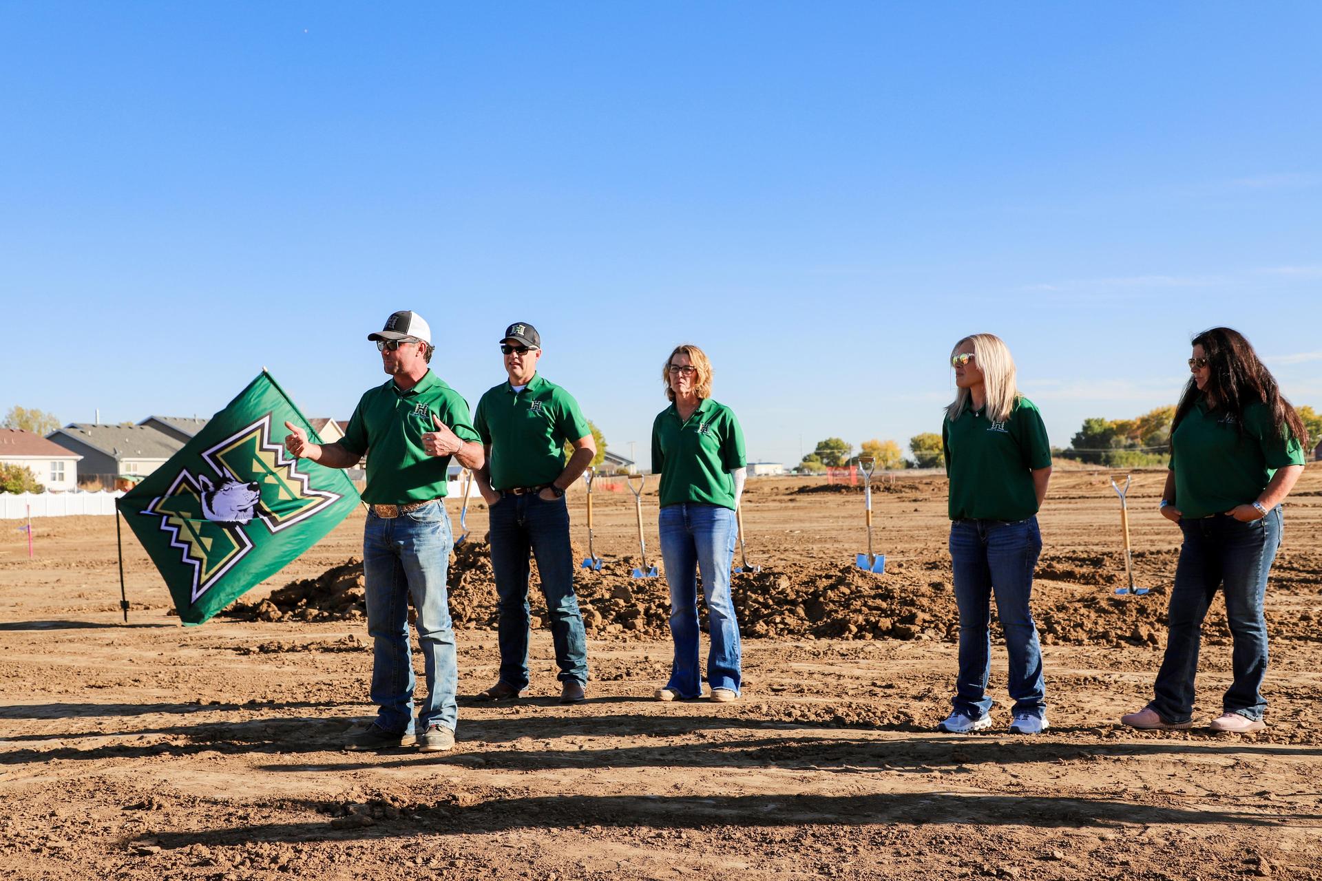 Adults in green shirts speaking at a construction site with a flag.