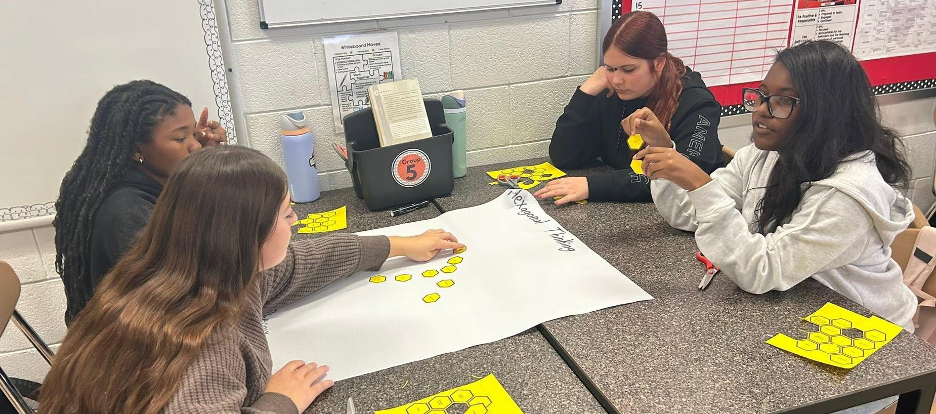 Four students collaborate at a table with worksheets and yellow cutouts.