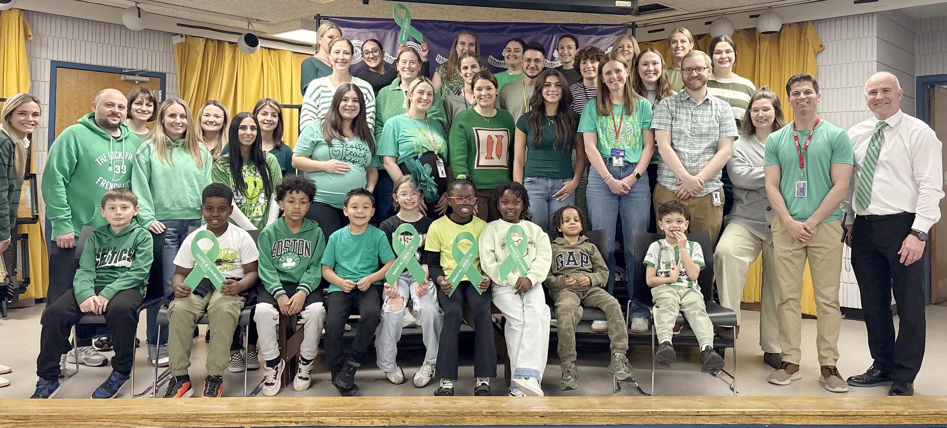 A large gathering of people in green clothing, smiling together in a school setting.