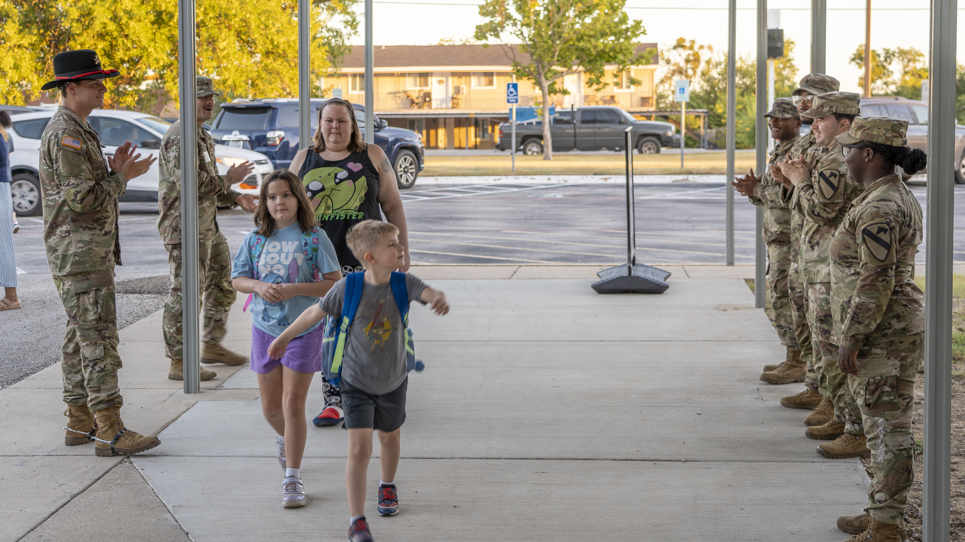 Children walking past smiling military personnel under a covered walkway.