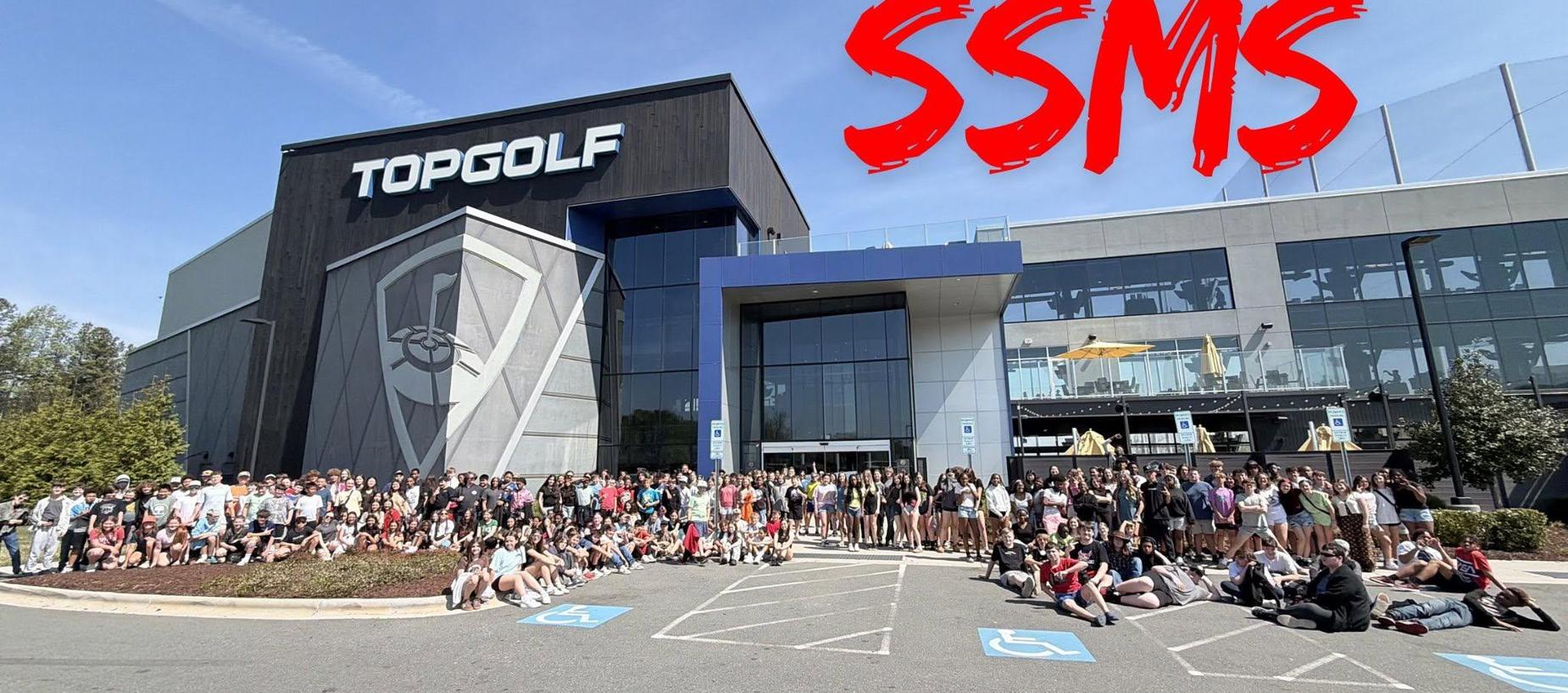 A large group of people gathered outside a Topgolf venue with a clear blue sky.