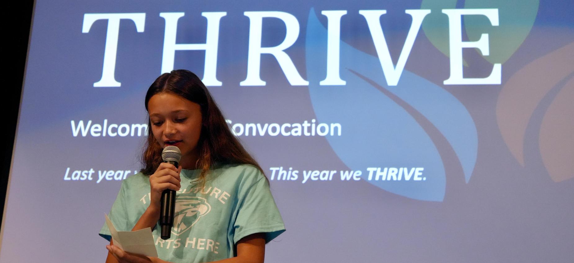 Girl reading from a paper on stage with a thrive backdrop.