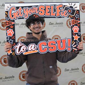 Smiling young man holding a colorful frame that says 'Get your SELFie to a CSU!'