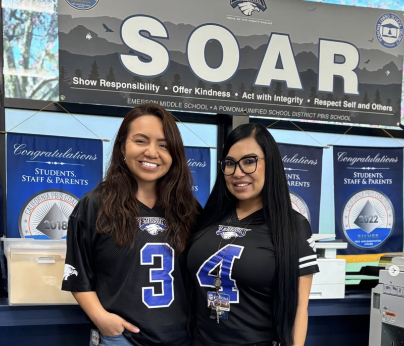 two teachers pose in the schools office with a big banner that says SOAR
