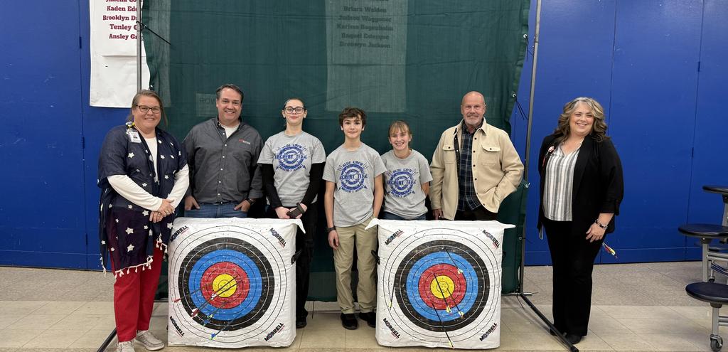 Students and adults smiling standing with archery arrows in targets on the ground