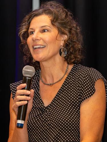 Woman speaking into a microphone, wearing a polka dot dress and earrings.