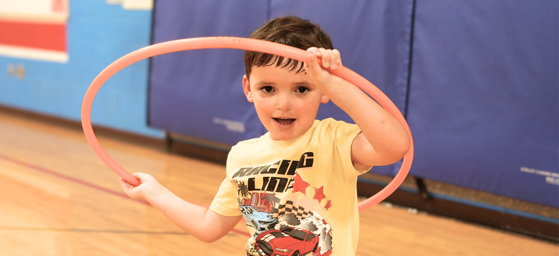 Boy holding a hula hoop excitedly in a gym setting.