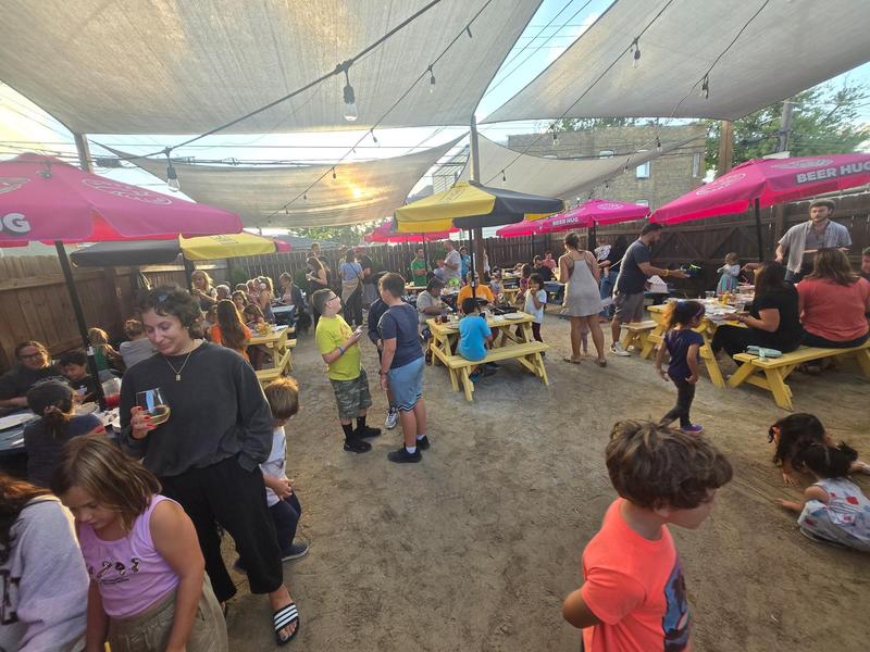 Busy outdoor gathering with families enjoying food under shaded picnic areas.