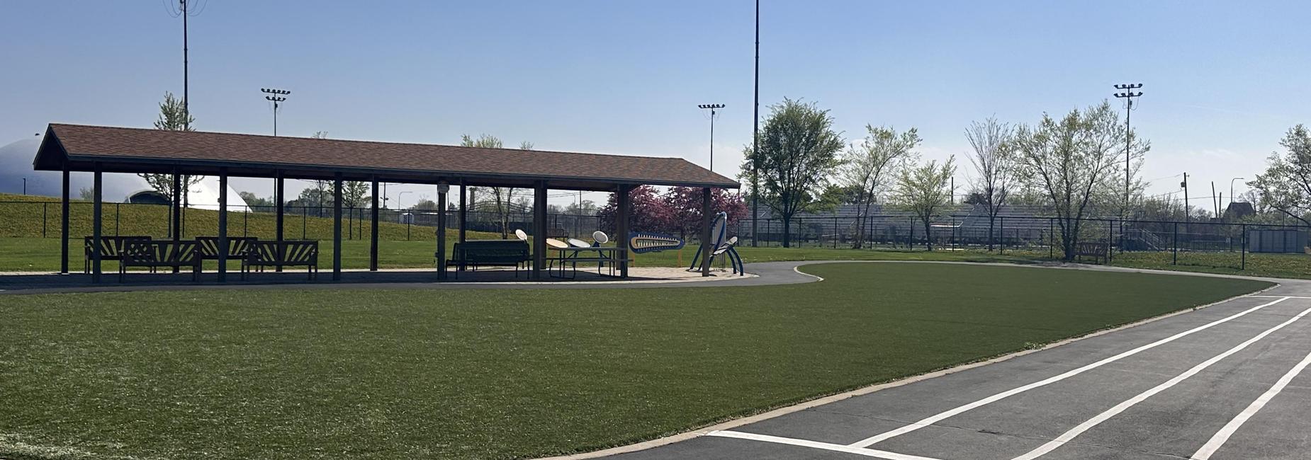 Open grassy area with a shelter and seating, surrounded by sports fields.