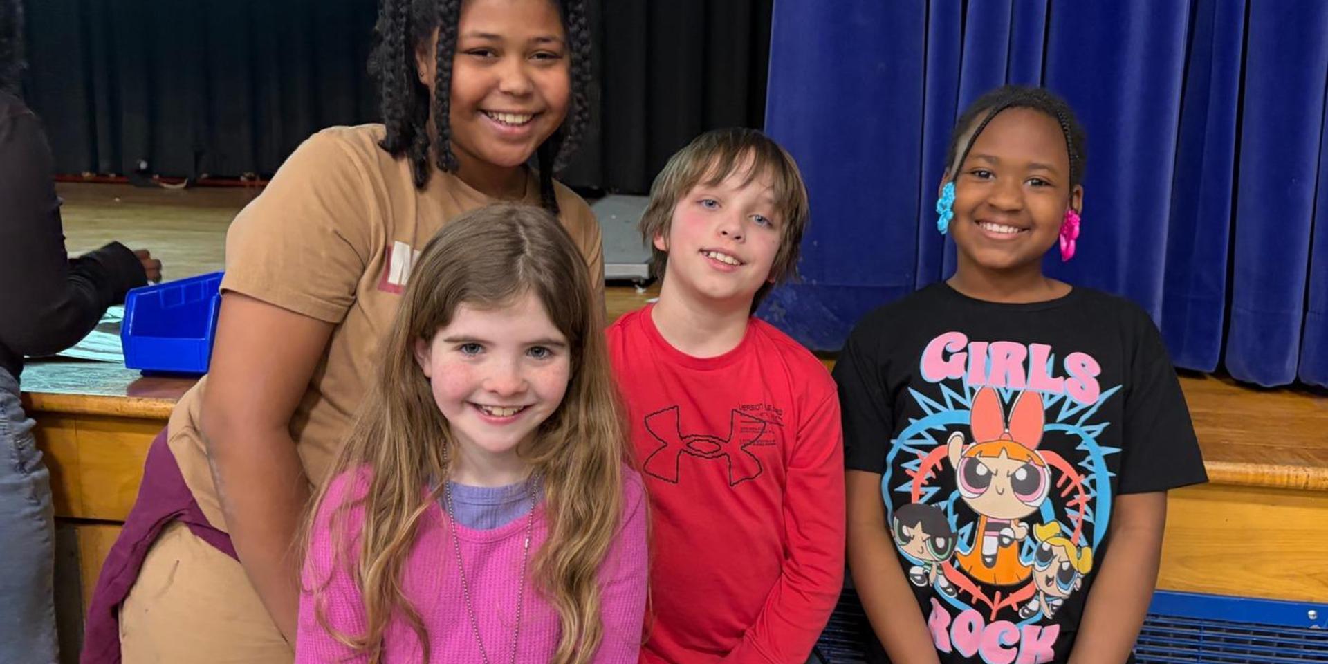 Four children smiling together on stage in a school auditorium.