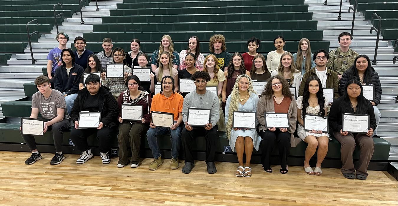 A group of students sitting on bleachers, holding certificates, smiling together.
