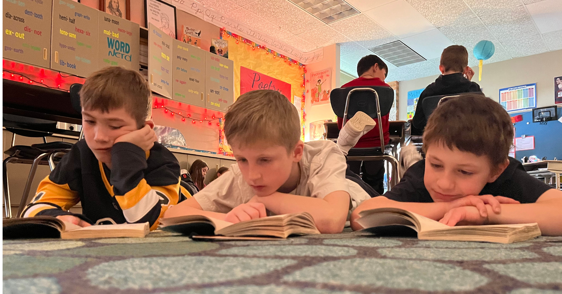 Three children lying on a carpet, reading books in a classroom setting.