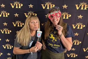 Two women pose in front of a black and gold “VIP” backdrop, holding playful props including a “First Day” sign and apple-shaped glasses.