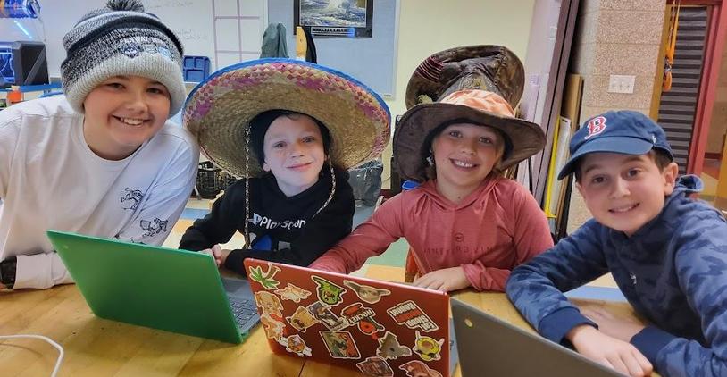 Four children wearing various hats sitting at a table with laptops.