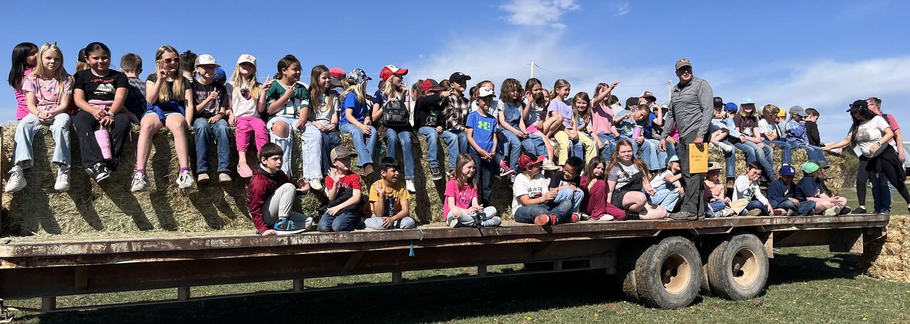 A large group of children sitting on hay bales in a sunny outdoor setting.