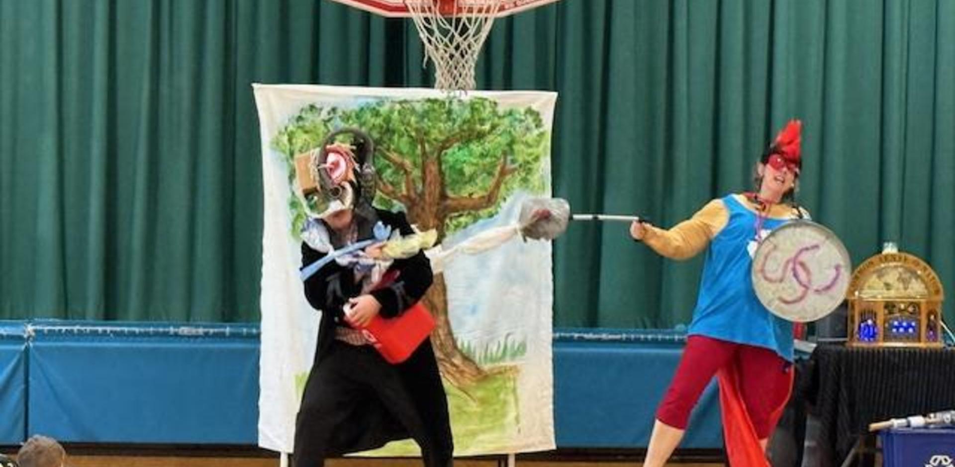 Two performers in costumes act out a comedic scene in front of a painted backdrop.