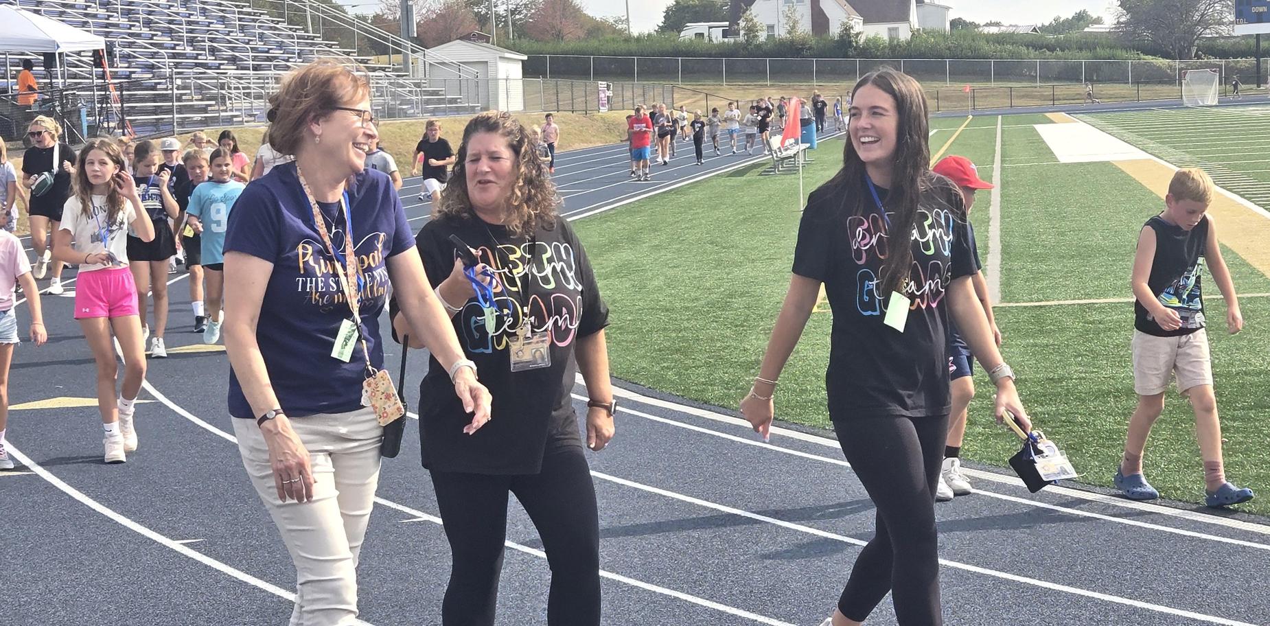 Three women walking and laughing on a track with kids in the background.