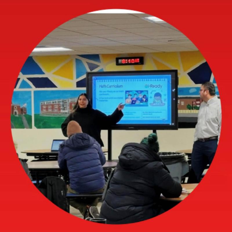 A woman in a classroom with adults, standing in front of a large screen